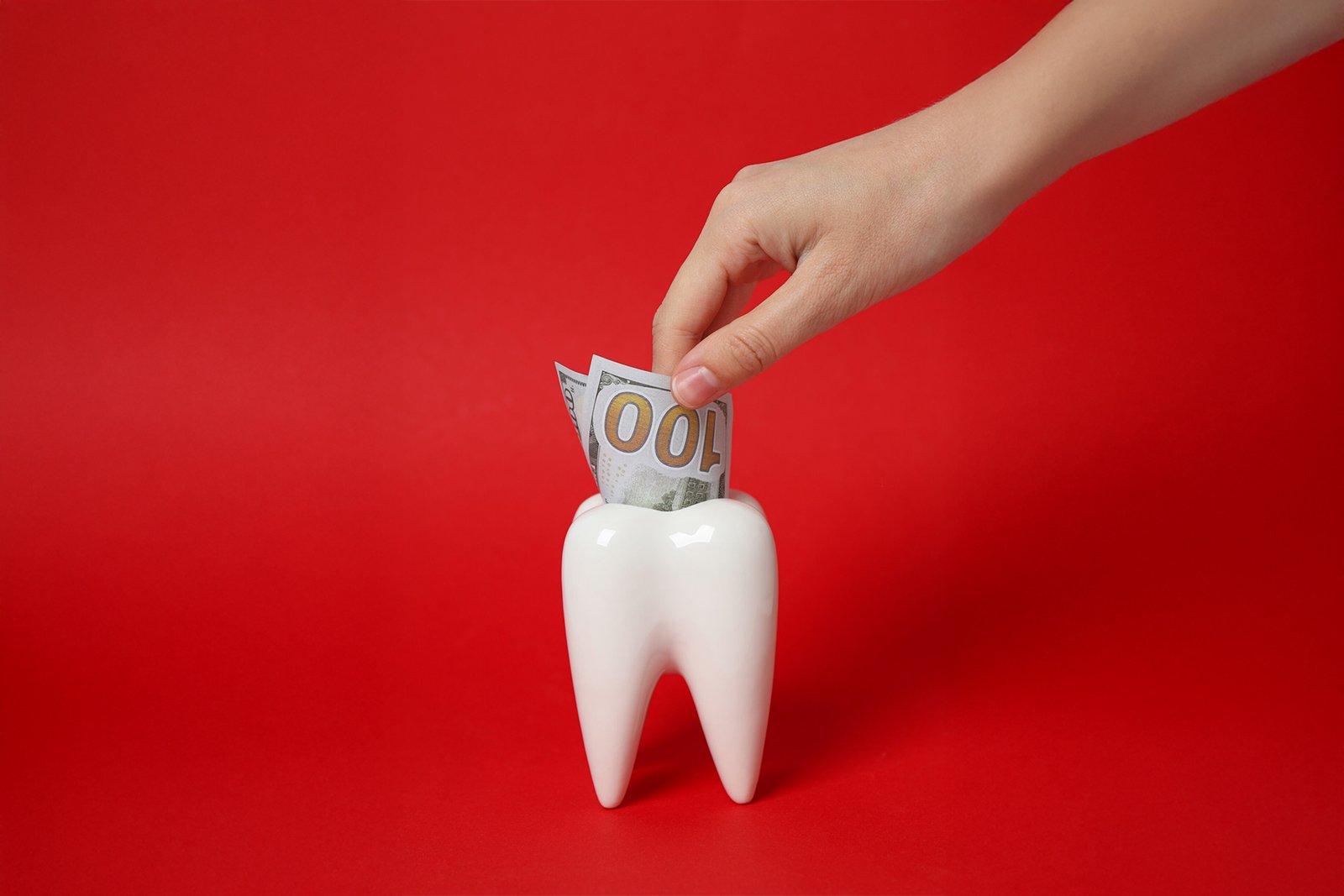 Hand placing money into a tooth-shaped container on red background, symbolizing dental prices Turkey